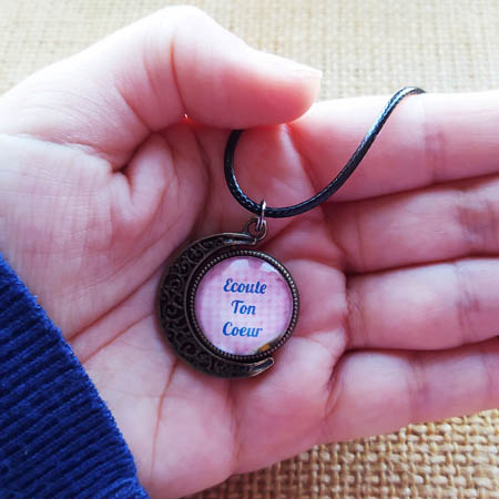 photographie d'une main qui tient un collier au pendentif eu forme de lune et au message inspirant Ecoute ton Coeur